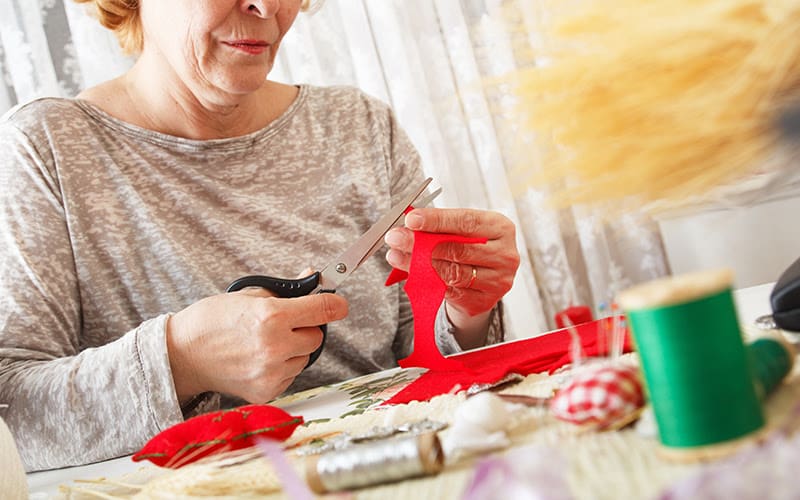 Senior women woman cuts red fabric with scissors, and making ornament.