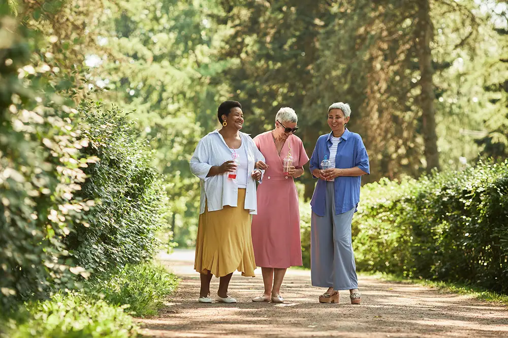 Three Sedgebrook residents stroll along a sunny, tree-lined outdoor path holding cold drinks and enjoying active, fulfilling retirement plans for individuals.