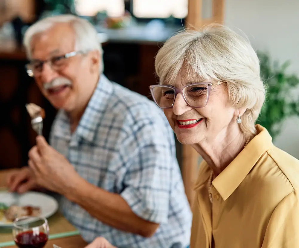 A senior woman wearing glasses smiles warmly during a meal while seated next her husband, showcasing the social dining experience enjoyed by seniors at Sedgebrook with secure retirement plans.